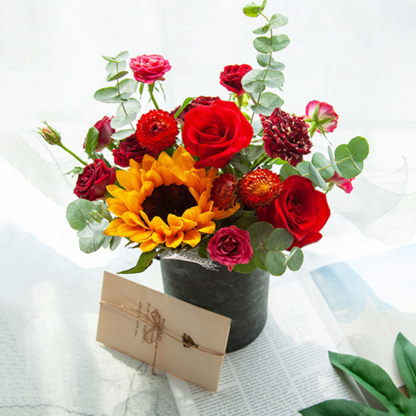 Medium assortment of sunflower, roses, lisianthus, and eucalyptus arranged in a cylinder gift box