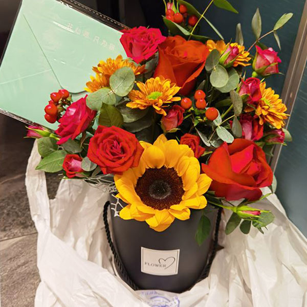 Medium assortment of sunflower, roses, lisianthus, and eucalyptus arranged in a cylinder gift box
